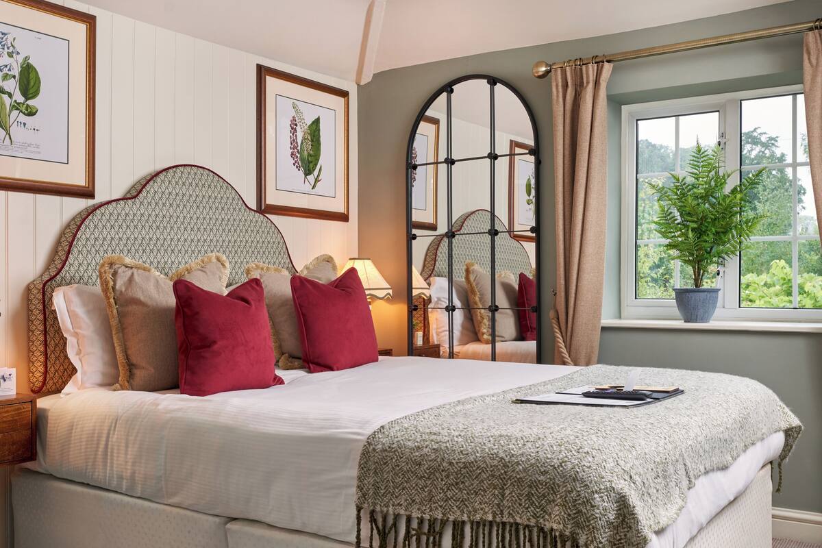 A neatly made bed with a headboard, a bedside lamp, and a window with a view of greenery.