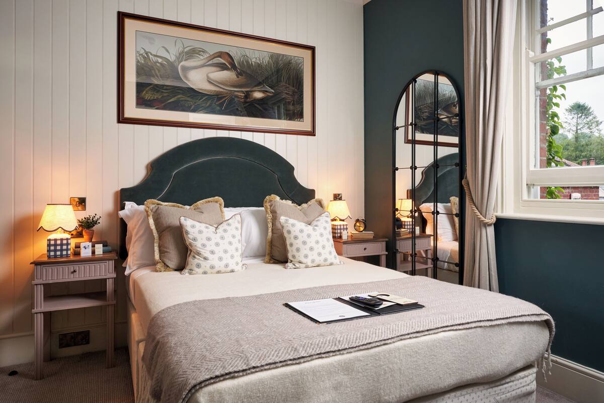 A neatly made bed with a headboard, two bedside tables with lamps, a framed picture on the wall, and a window with curtains.