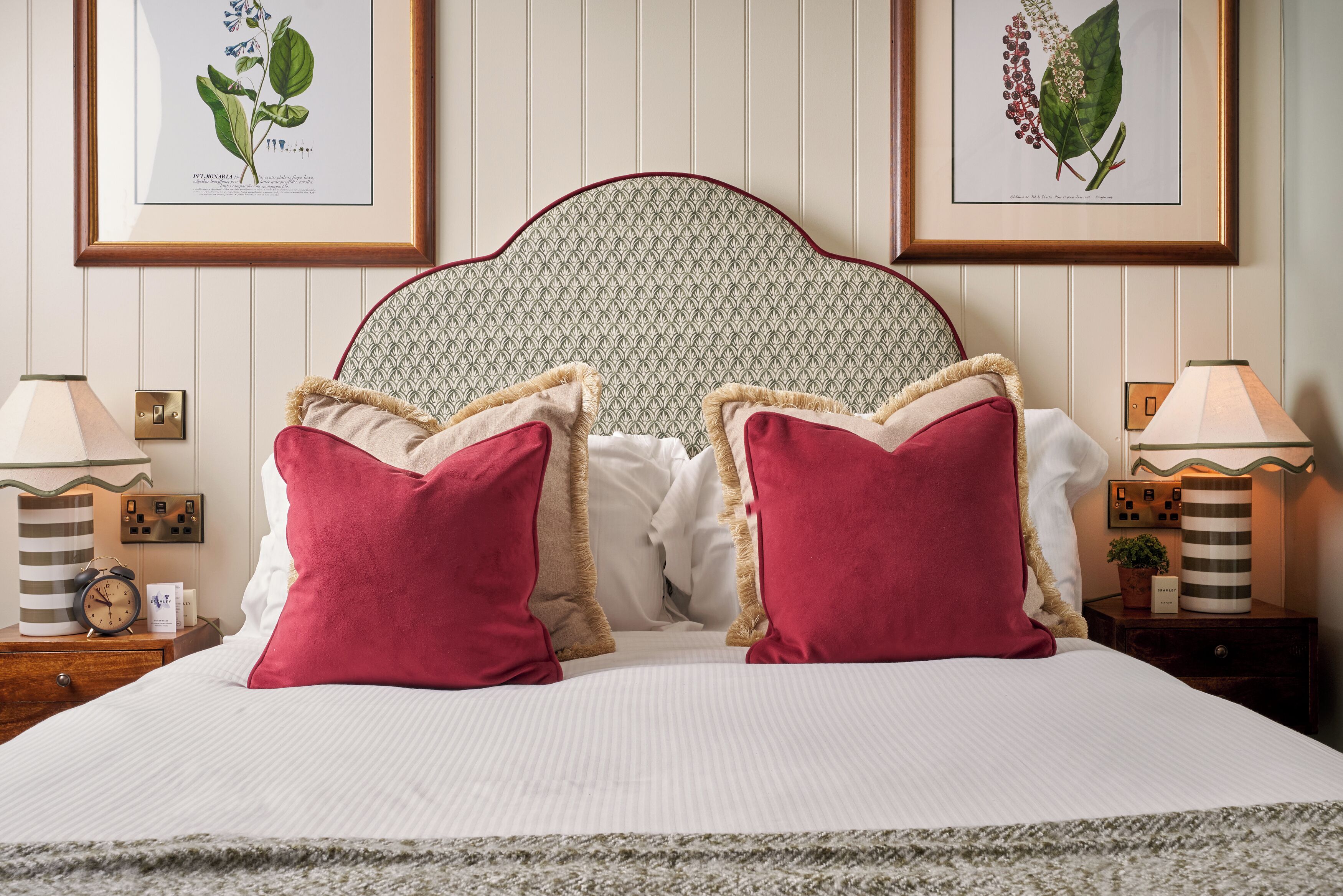 A neatly made bed with two red pillows, a headboard with a patterned design, and two framed botanical illustrations on the wall.