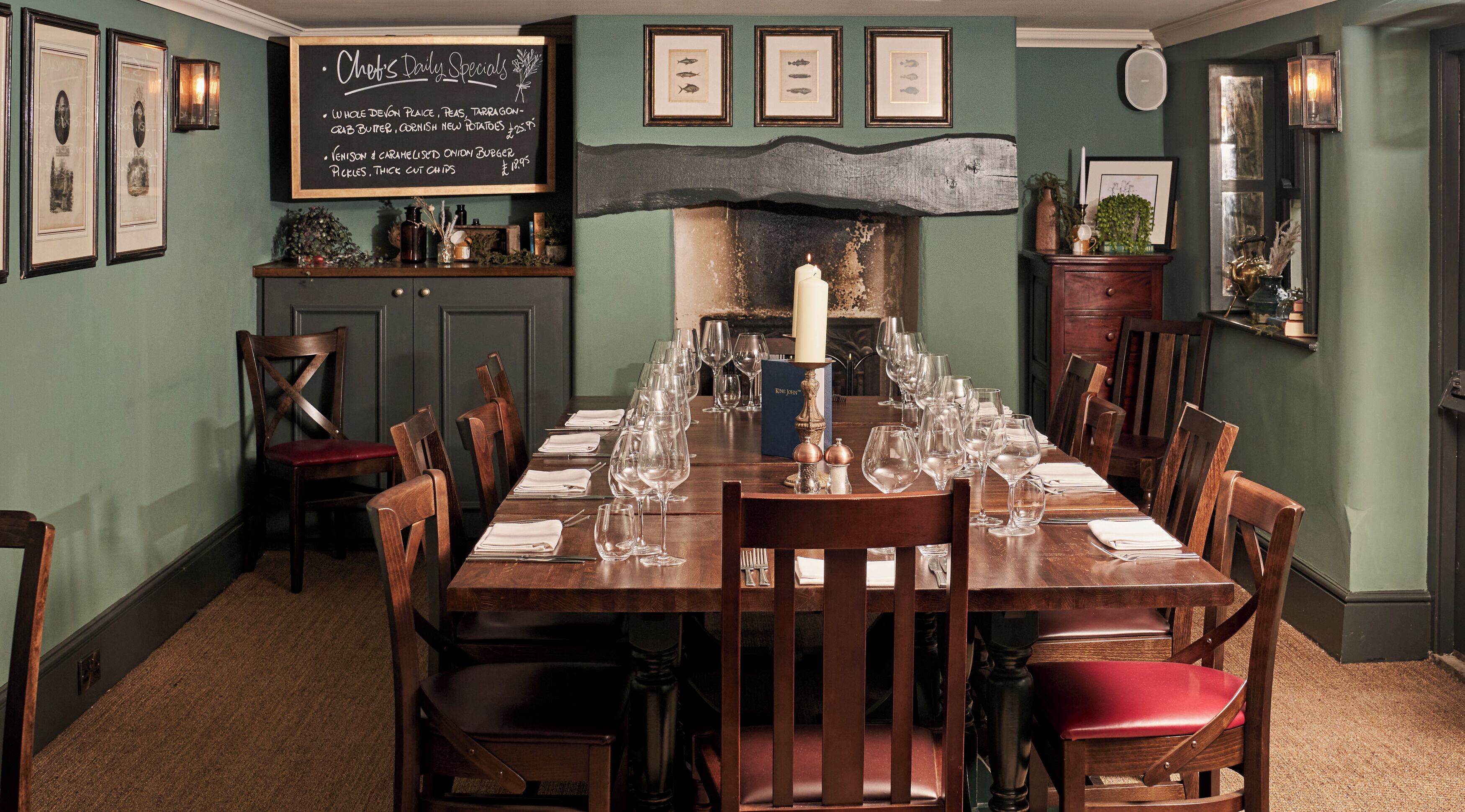 A dining room with a long wooden table set for a meal, framed pictures on the walls, and a chalkboard menu.
