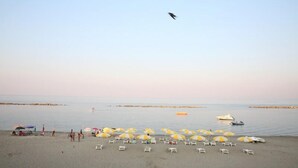 Uma praia com espreguiçadeiras e guarda-sóis, um barco e uma pessoa a surfar.