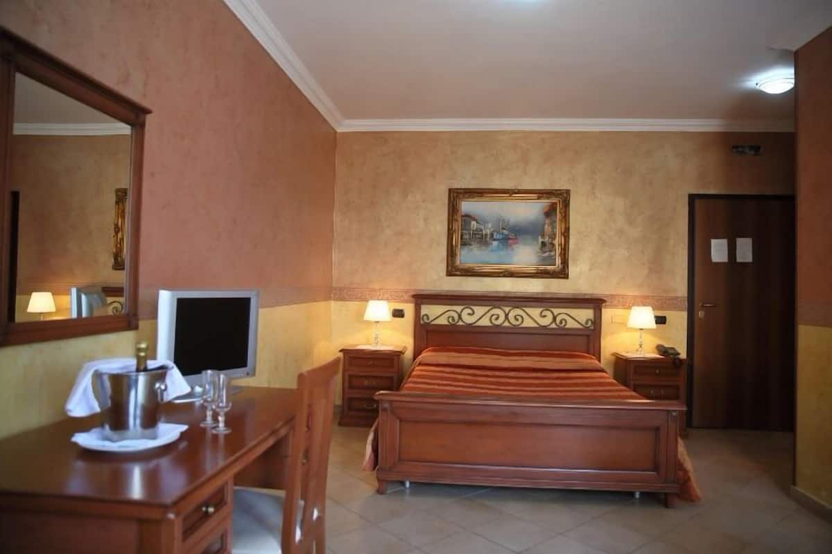 A hotel room with a bed, bedside tables, a desk with a computer, a mirror, and a painting on the wall.