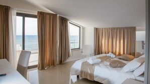 Quarto de hotel com uma cama grande, uma escrivaninha e uma cadeira. Vê-se o oceano pelas janelas.