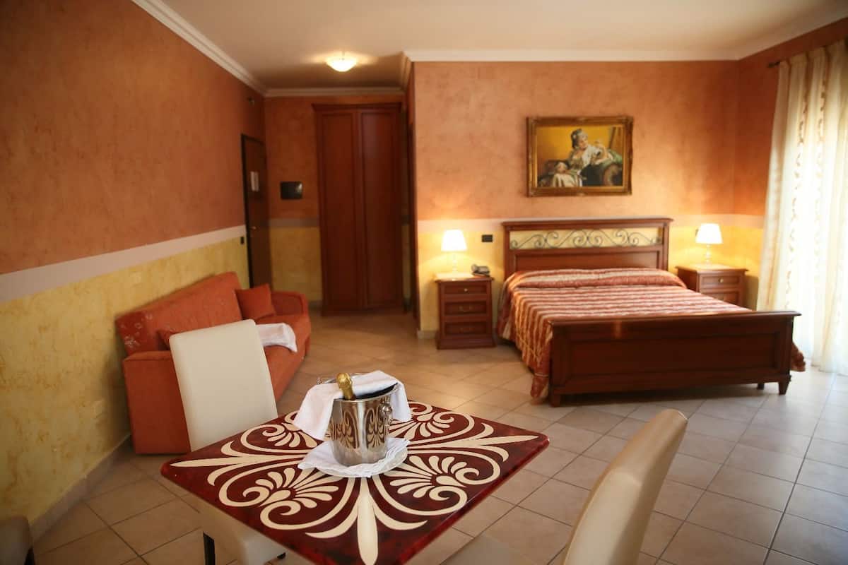 A hotel room with a bed, bedside tables, a table with a decorative table runner, a sofa, and a painting on the wall.