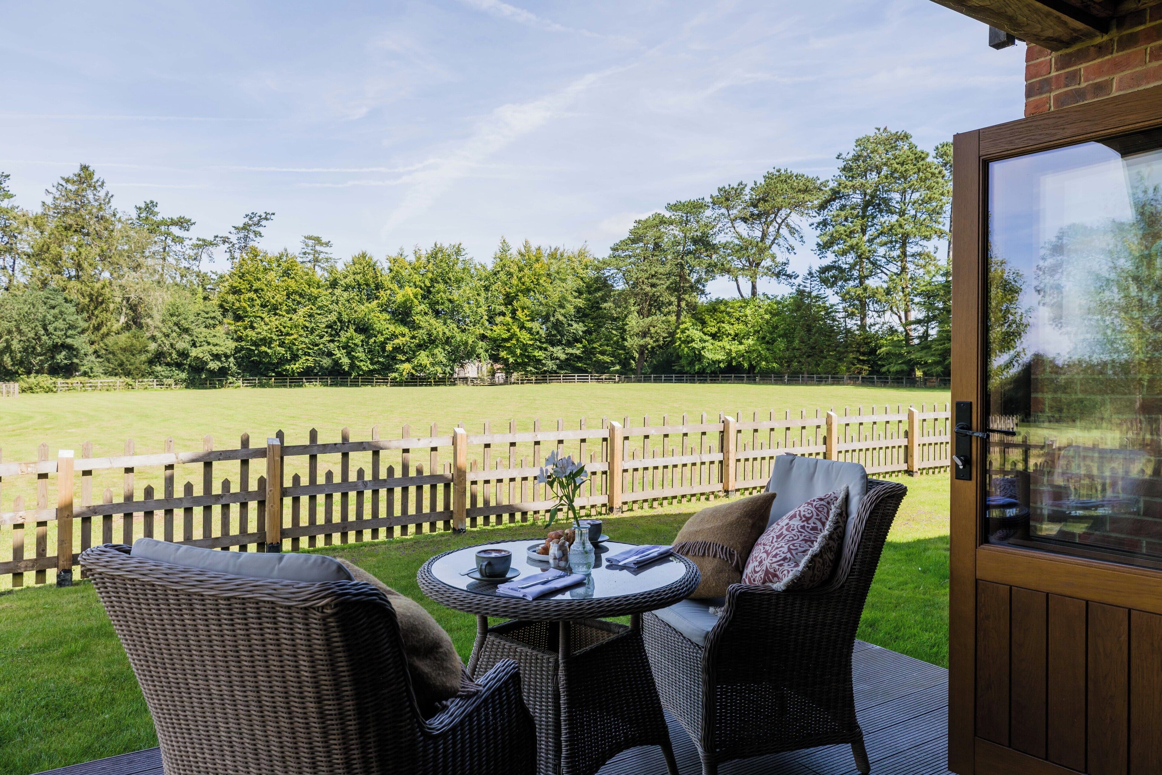 An outdoor seating area with wicker furniture and a table, overlooking a grassy field and wooden fence.