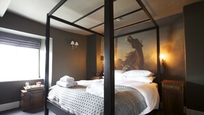 A four-poster bed with a grey blanket, a wooden nightstand with a lamp, and a painting on the wall. - The Kings Arms (Badminton)