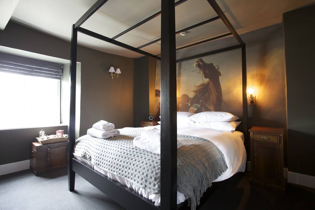 A four-poster bed with a grey blanket, a wooden nightstand with a lamp, and a painting on the wall.