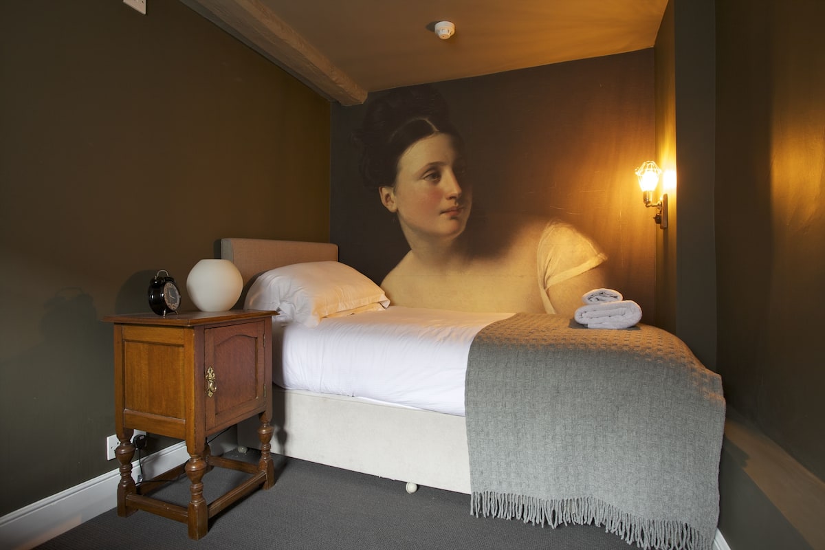 A bedroom with a bed, a nightstand, a wall-mounted lamp, and a painting.