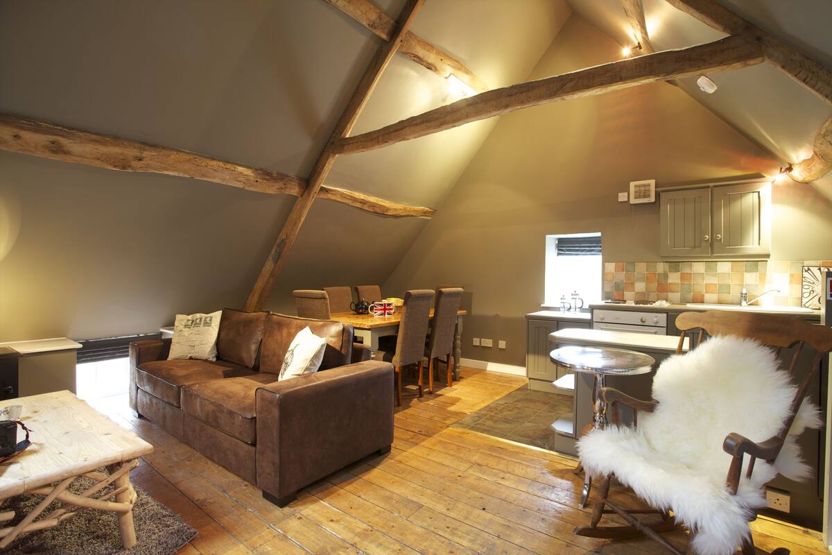 A cozy loft-style living area with a wooden ceiling, a brown leather sofa, a fluffy white chair, and a dining table with chairs.