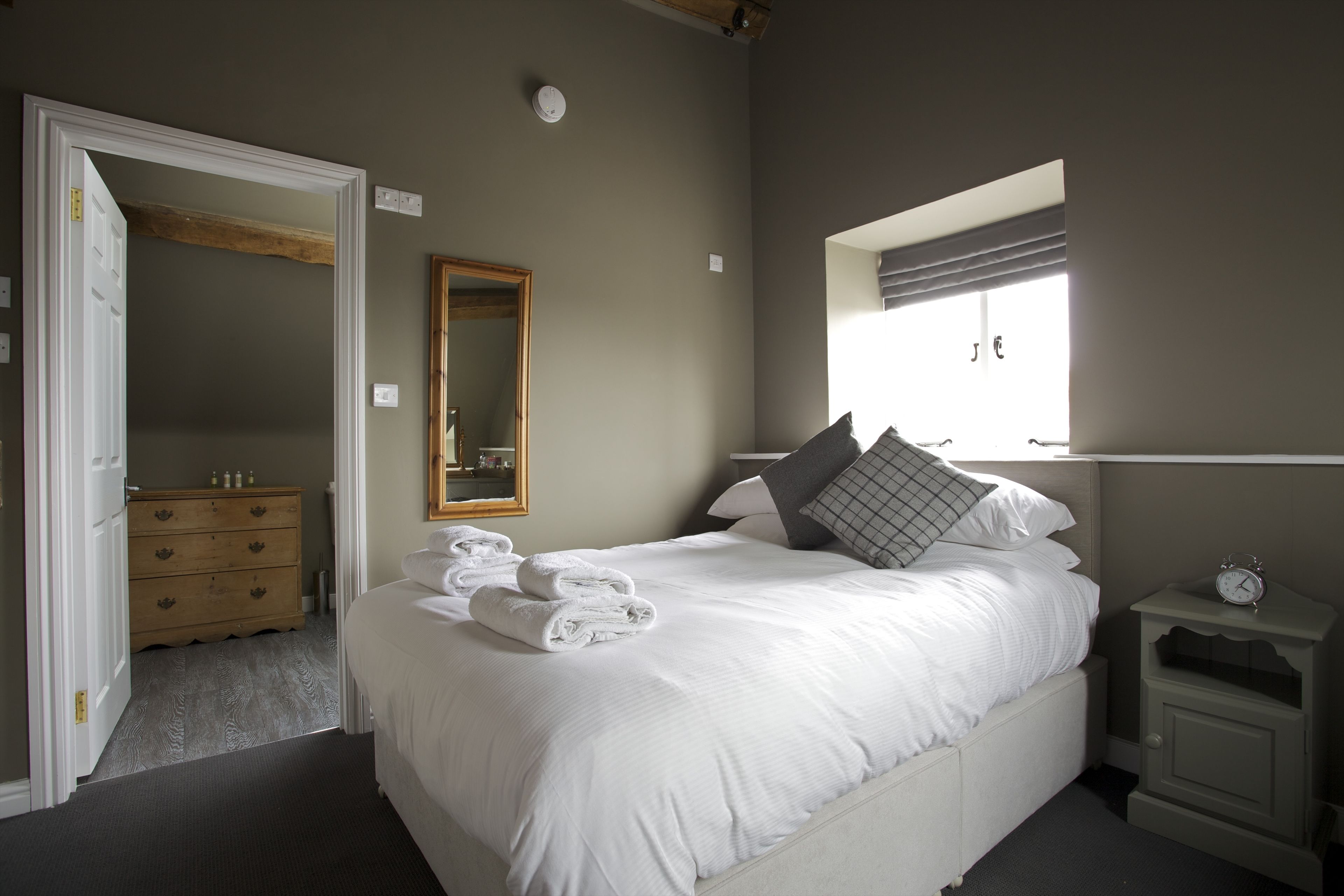 Una cama bien hecha con almohadas y toallas dobladas, una mesita de noche con un reloj, una ventana con persianas y un aparador de madera al fondo.