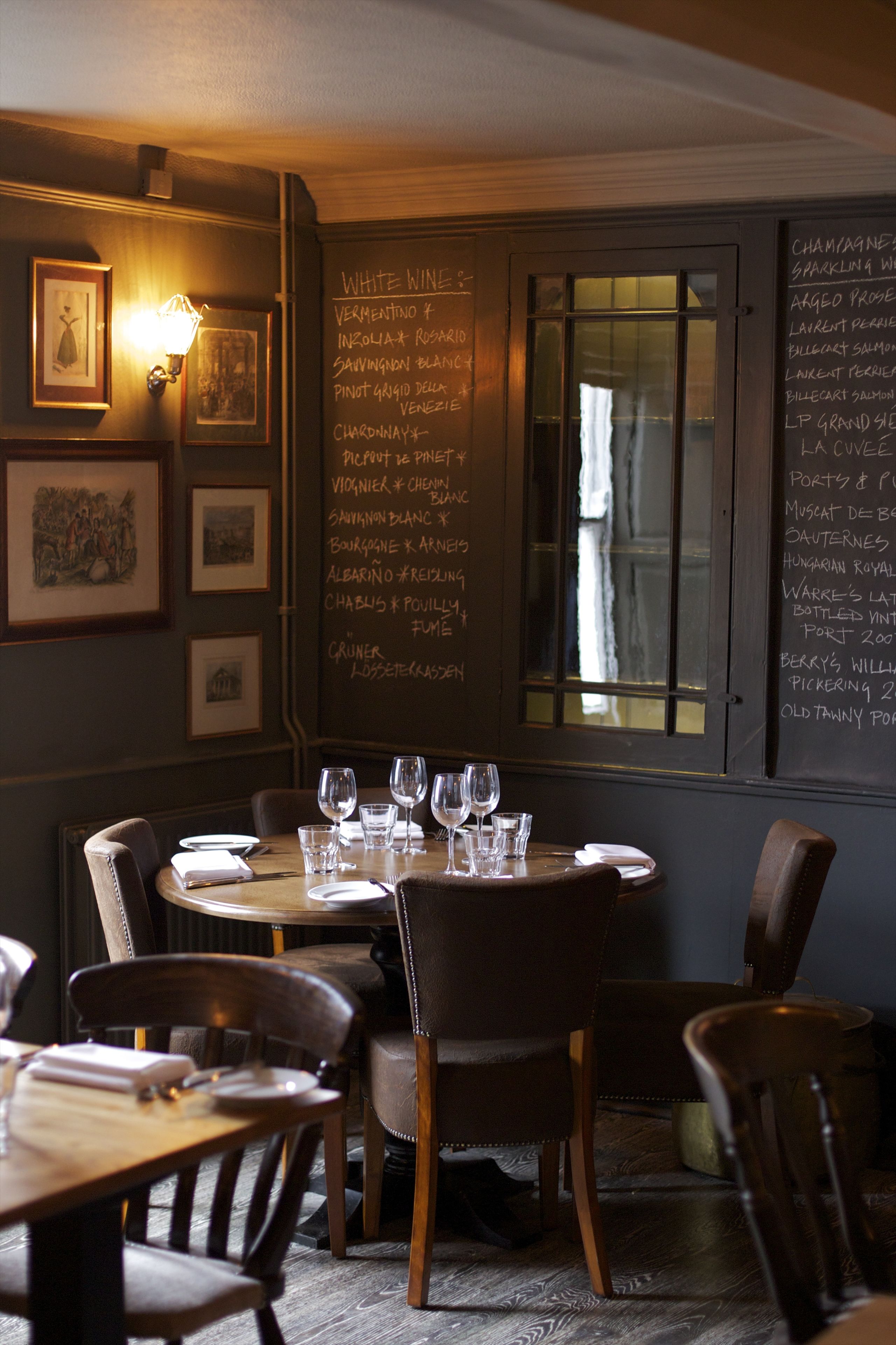 A restaurant interior with a chalkboard menu listing white wines and other beverages, framed pictures on the wall, and a table set for dining.