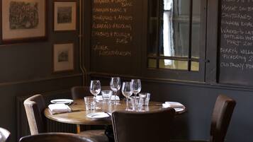 Interior de un restaurante con un menú en pizarra que enumera vinos blancos y otras bebidas, cuadros en la pared y una mesa lista para comer.