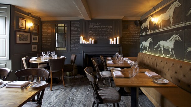 A restaurant with wooden tables and chairs, a fireplace, and wall-mounted candles. - The Kings Arms (Badminton)