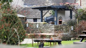 A rustic outdoor seating area with stone walls, a wooden table, and metal chairs. - The Kings Arms (Badminton)