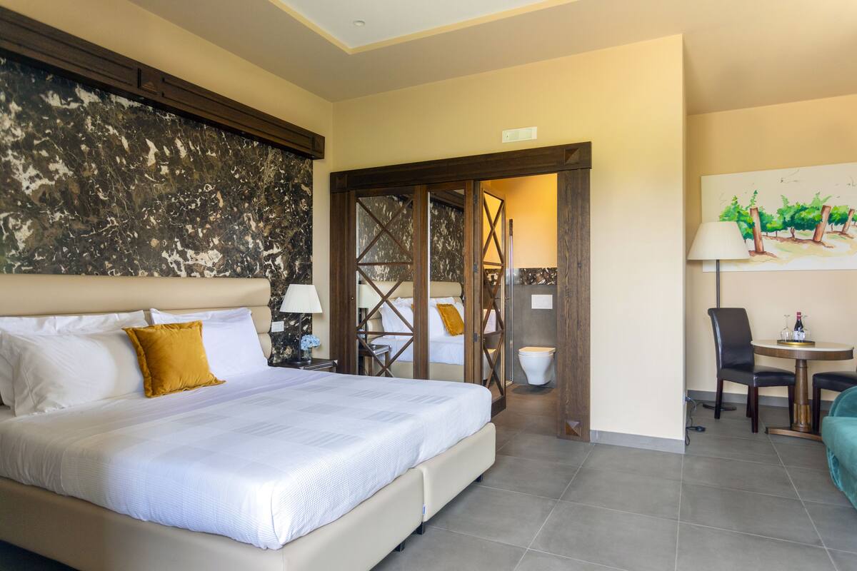A hotel room with a large bed, a wooden door, a dining table with chairs, and a painting on the wall.