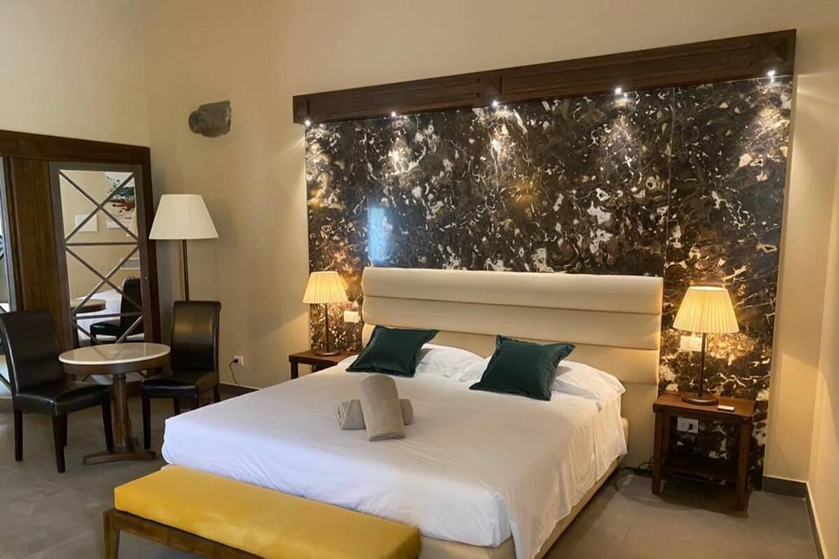 A hotel room with a large bed, two bedside tables with lamps, a bench, a chair, and a decorative wall panel.