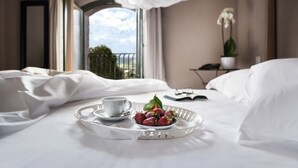 A hotel room with a bed, a tray with strawberries and a cup, a window with a view, and a plant on a side table. - Cavanera Etnea Resort & Wine Experience (Castiglione di Sicilia)