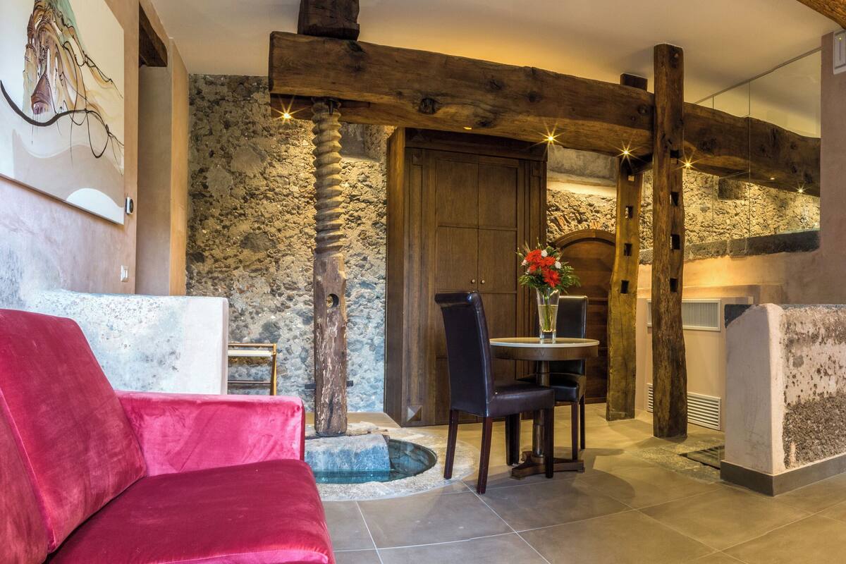 A modern hotel room with a stone wall, wooden beams, a bed, a red chair, and a dining table with chairs.