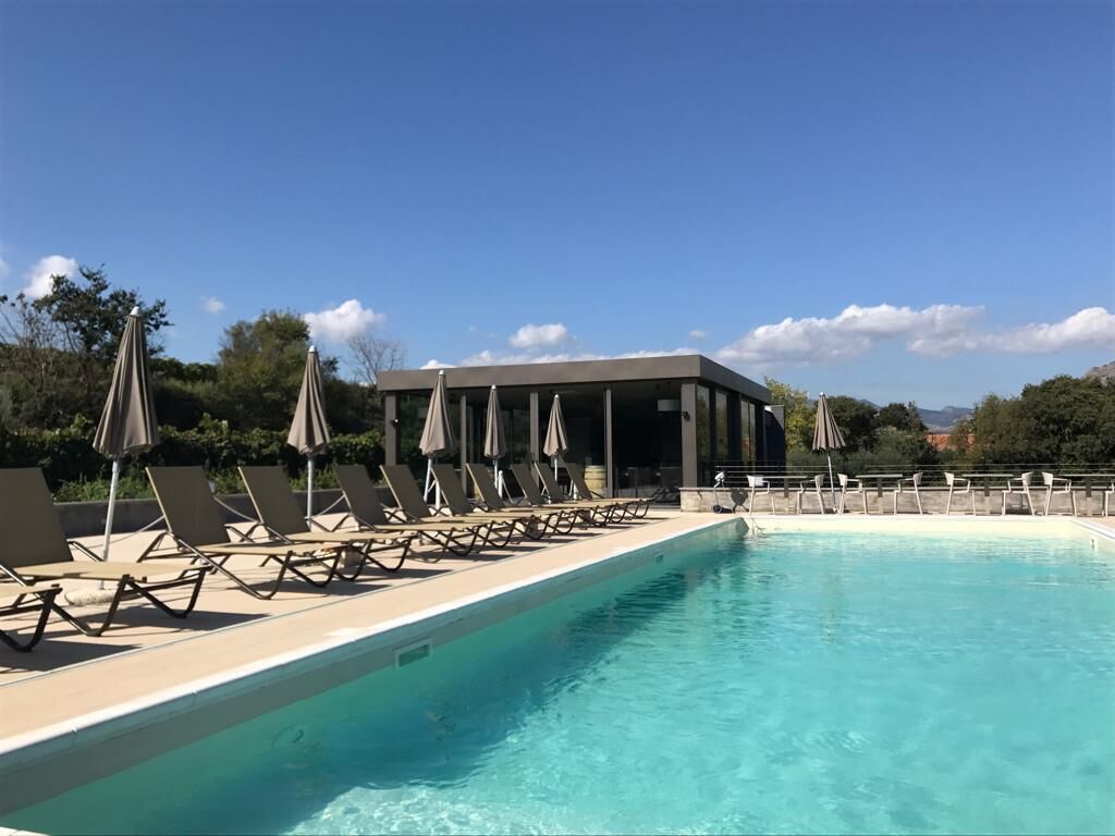 Ein Schwimmbecken mit Liegestühlen und Sonnenschirmen, umgeben von einem Gebäude mit einer überdachten Terrasse.