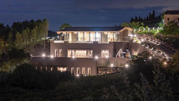 A modern building with multiple illuminated windows and a stone facade, surrounded by greenery and a sloped landscape. - Cavanera Etnea Resort & Wine Experience (Castiglione di Sicilia)
