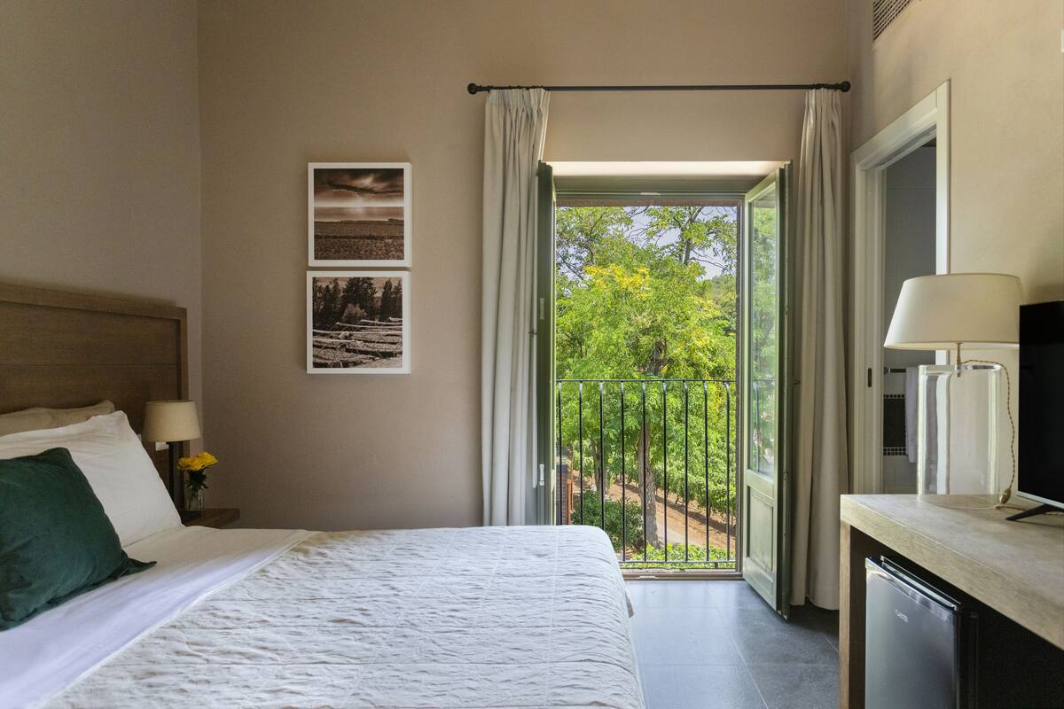 A hotel room with a bed, a desk with a lamp, a TV, and a balcony with curtains.