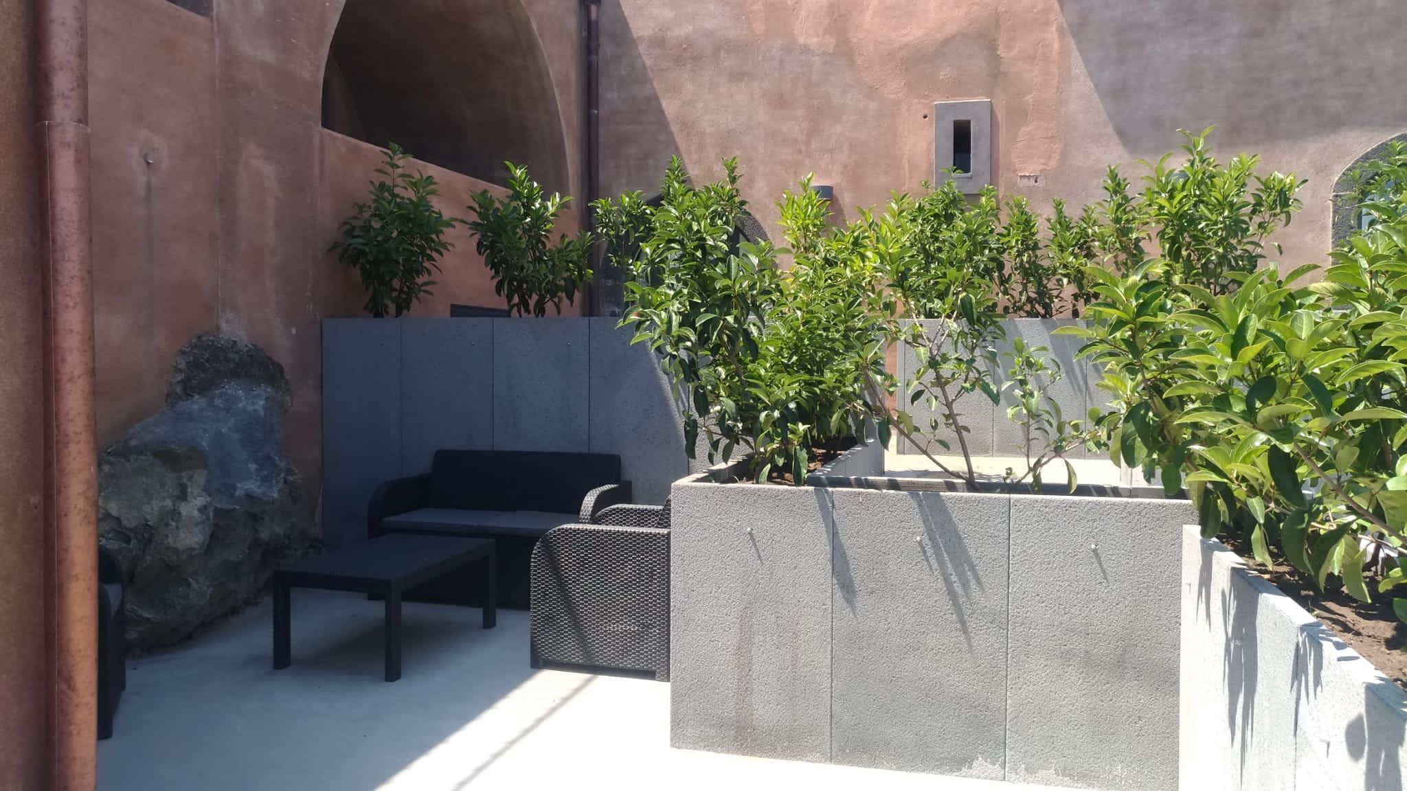 An outdoor seating area with a sofa and chairs, surrounded by planters with green shrubs.