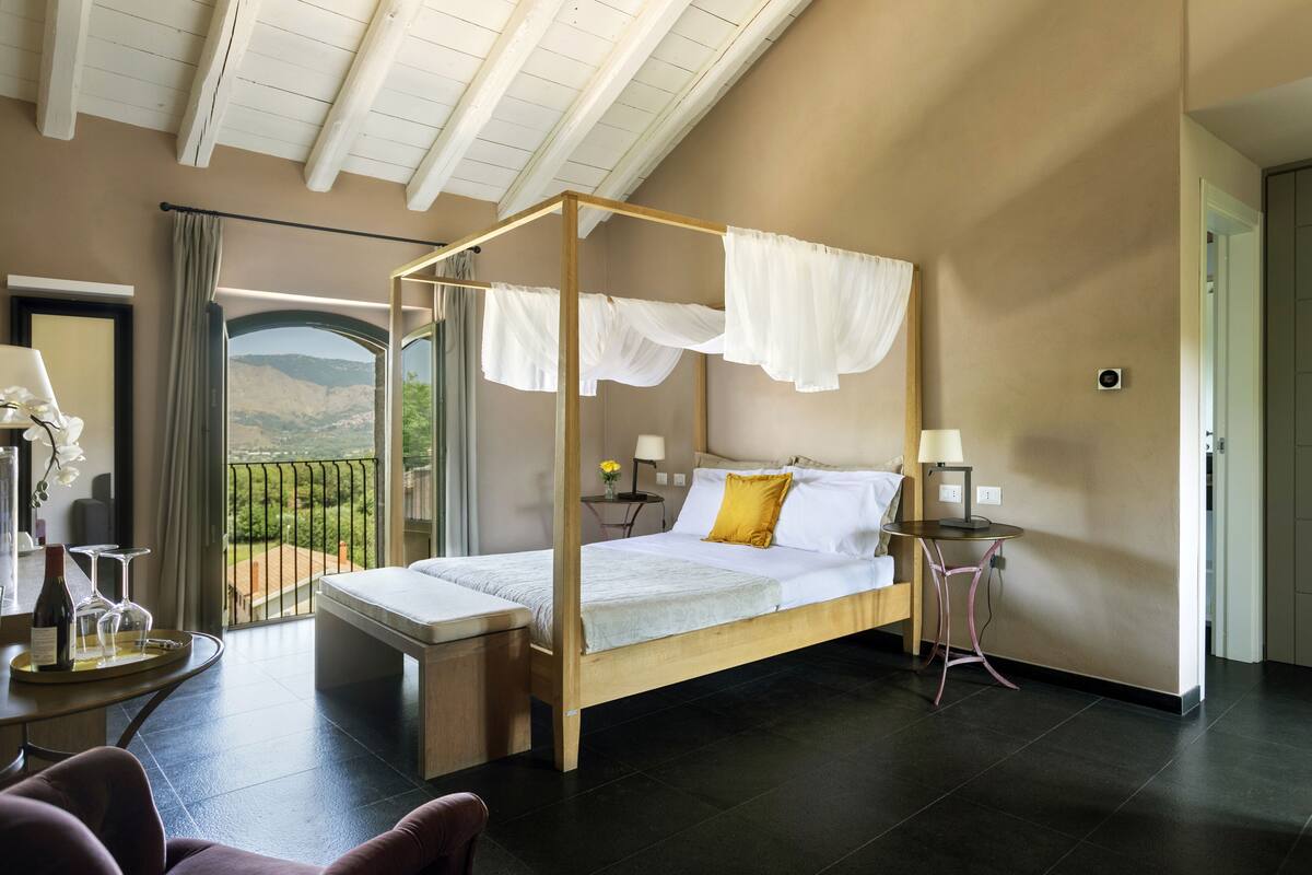 A four-poster bed with a view of the outdoors, a bedside table with a lamp, and a small table with a bottle and glasses.