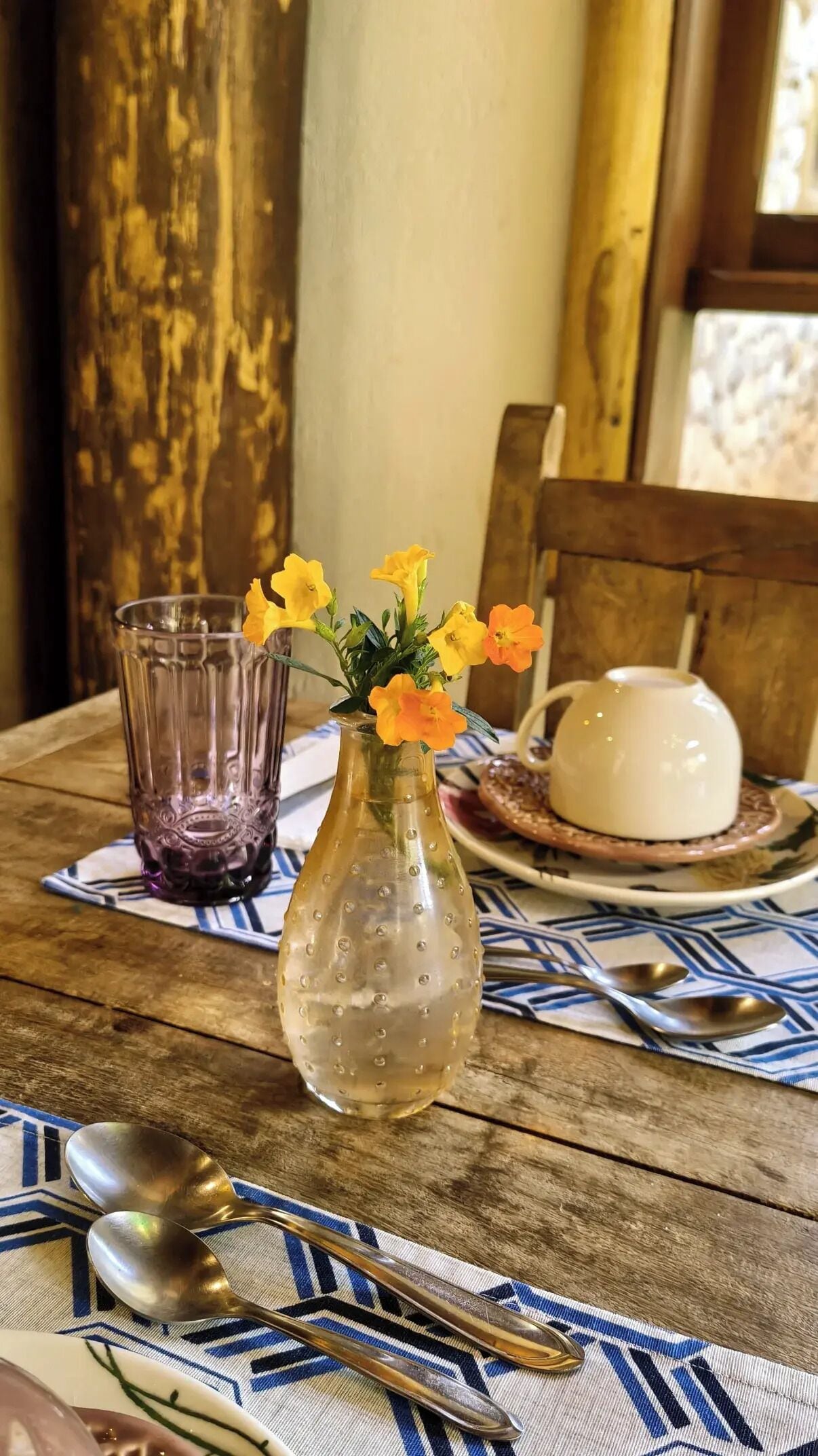 Una mesa de madera con un jarrón de vidrio transparente que contiene flores amarillas, una tetera blanca y dos cucharas sobre un mantel estampado.