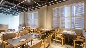 A dining area with multiple tables and chairs, large windows with blinds, and a ceiling with exposed pipes and lighting fixtures. - Hotel Mahadev Residency (Bhiwandi)