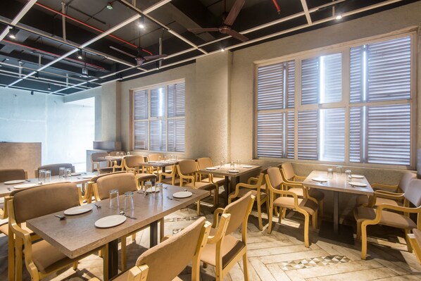 A dining area with multiple tables and chairs, large windows with blinds, and a ceiling with exposed pipes and lighting fixtures.