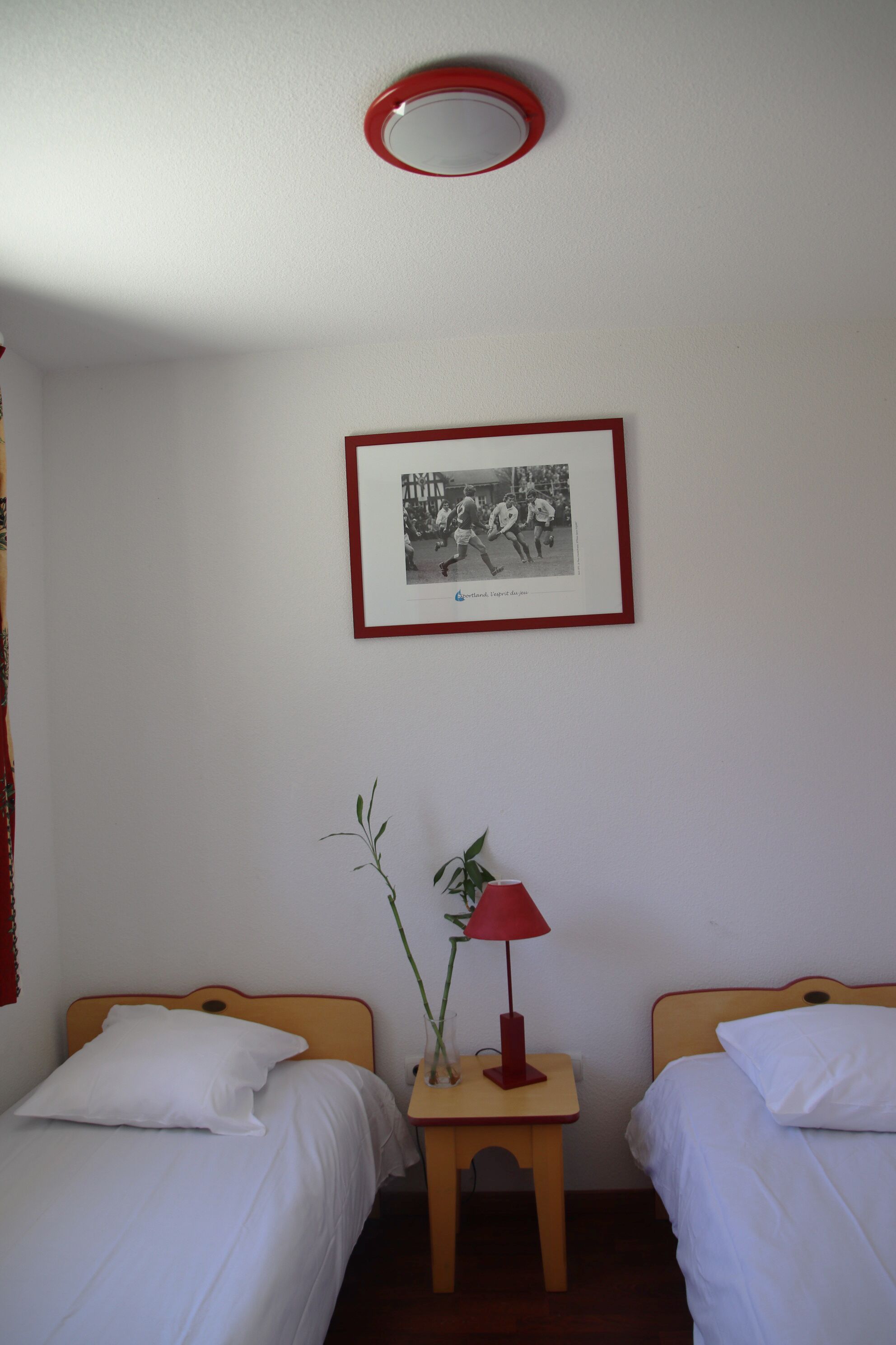 Une petite chambre propre, avec deux lits simples, un tableau encadré au mur, une petite table avec une lampe et un luminaire rouge au plafond.