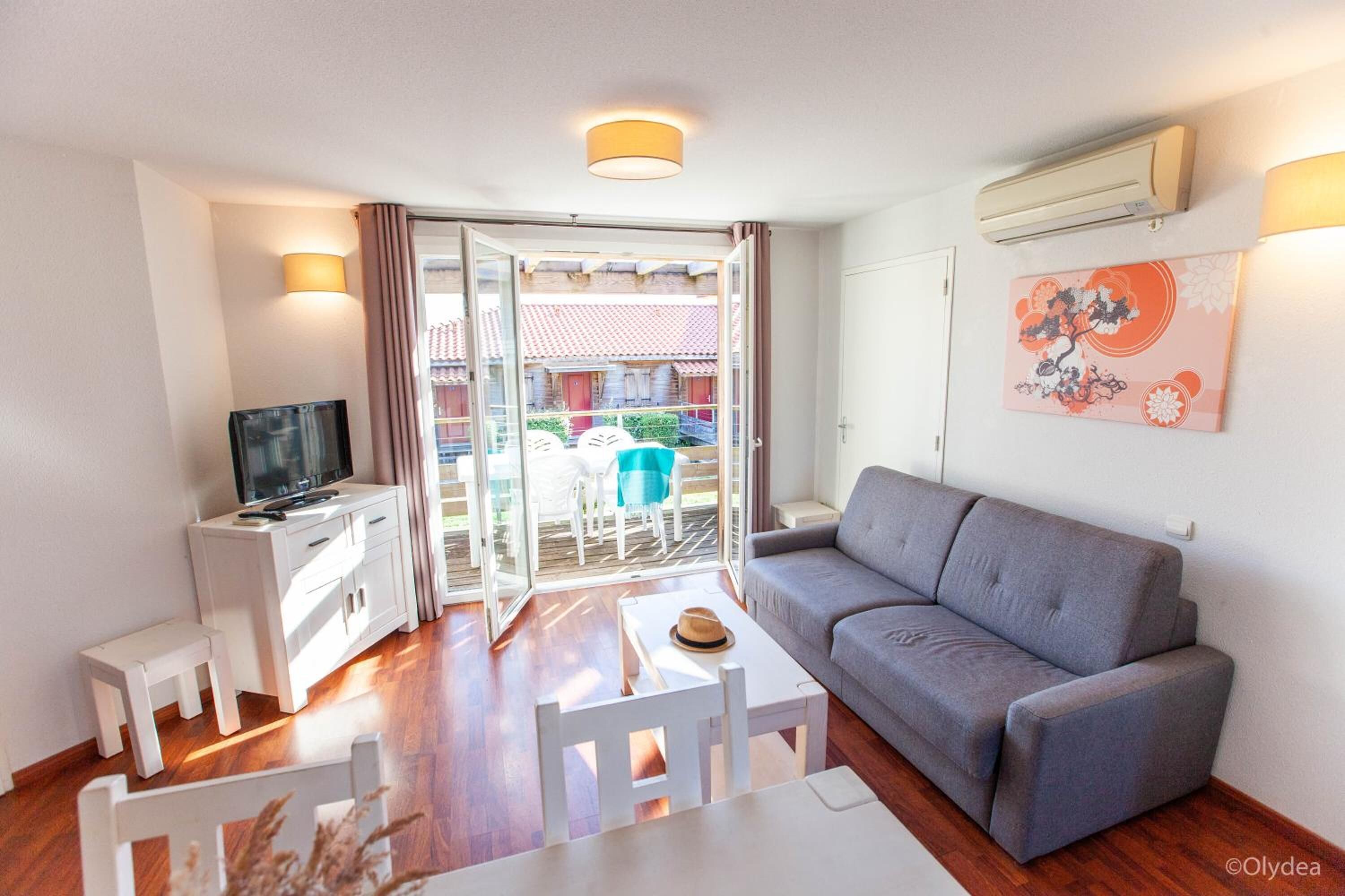 Un salón con un sofá gris, un televisor sobre un mueble blanco, una mesita con un sombrero y un balcón con mesa y sillas.