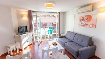 Un salón con un sofá gris, un televisor sobre un mueble blanco, una mesita con un sombrero y un balcón con mesa y sillas.