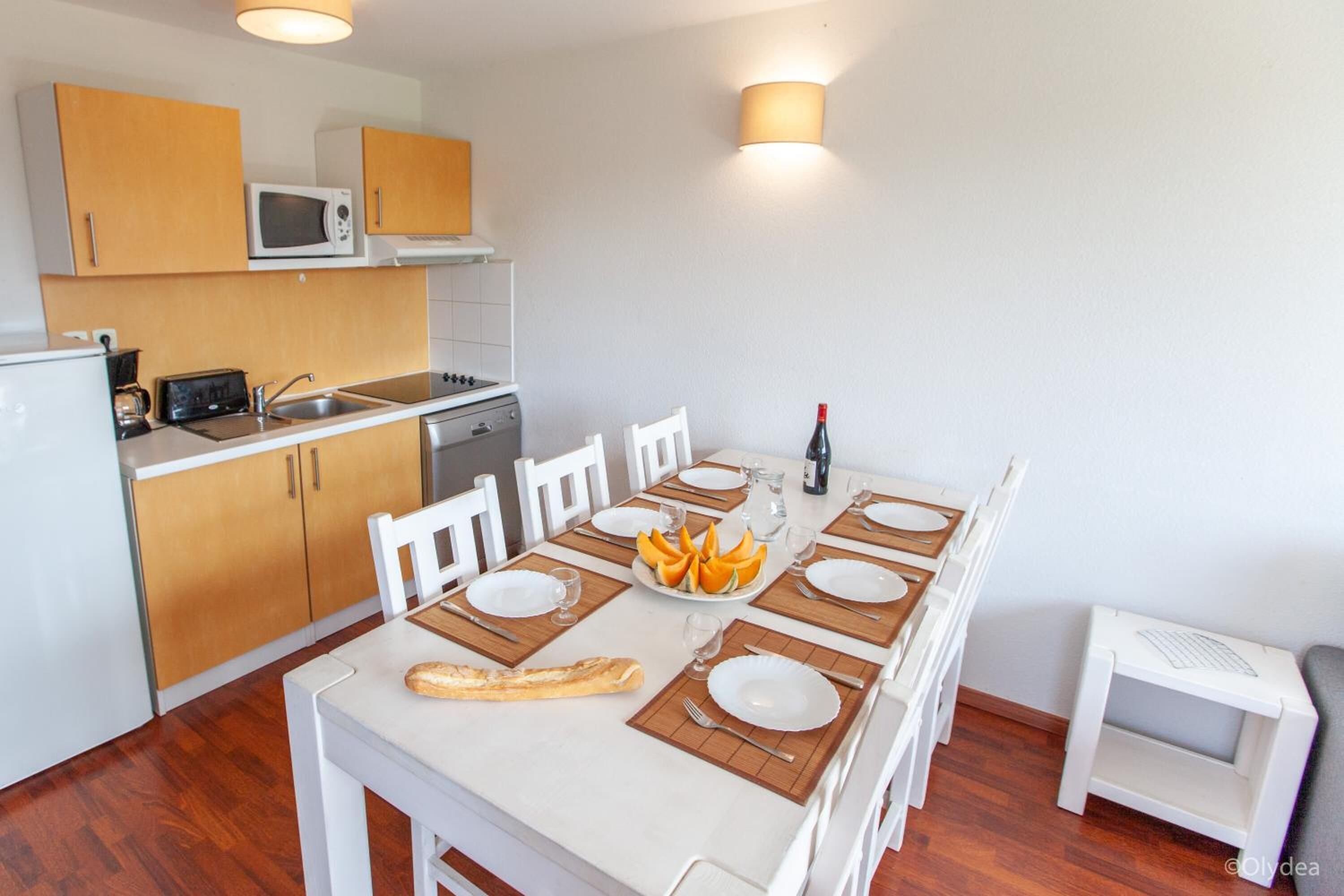 A compact kitchen with wooden cabinets, a microwave, and a dining table set for four with plates, cutlery, and a baguette.