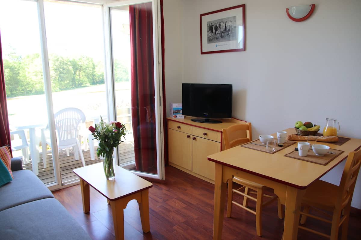 A living room with a sofa, a table with a vase of flowers, a television, and a dining area with chairs and a table set for breakfast.