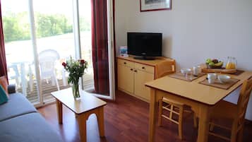 Un salón con sofá, una mesa con un jarrón de flores, un televisor y un comedor con sillas y una mesa preparada para el desayuno.