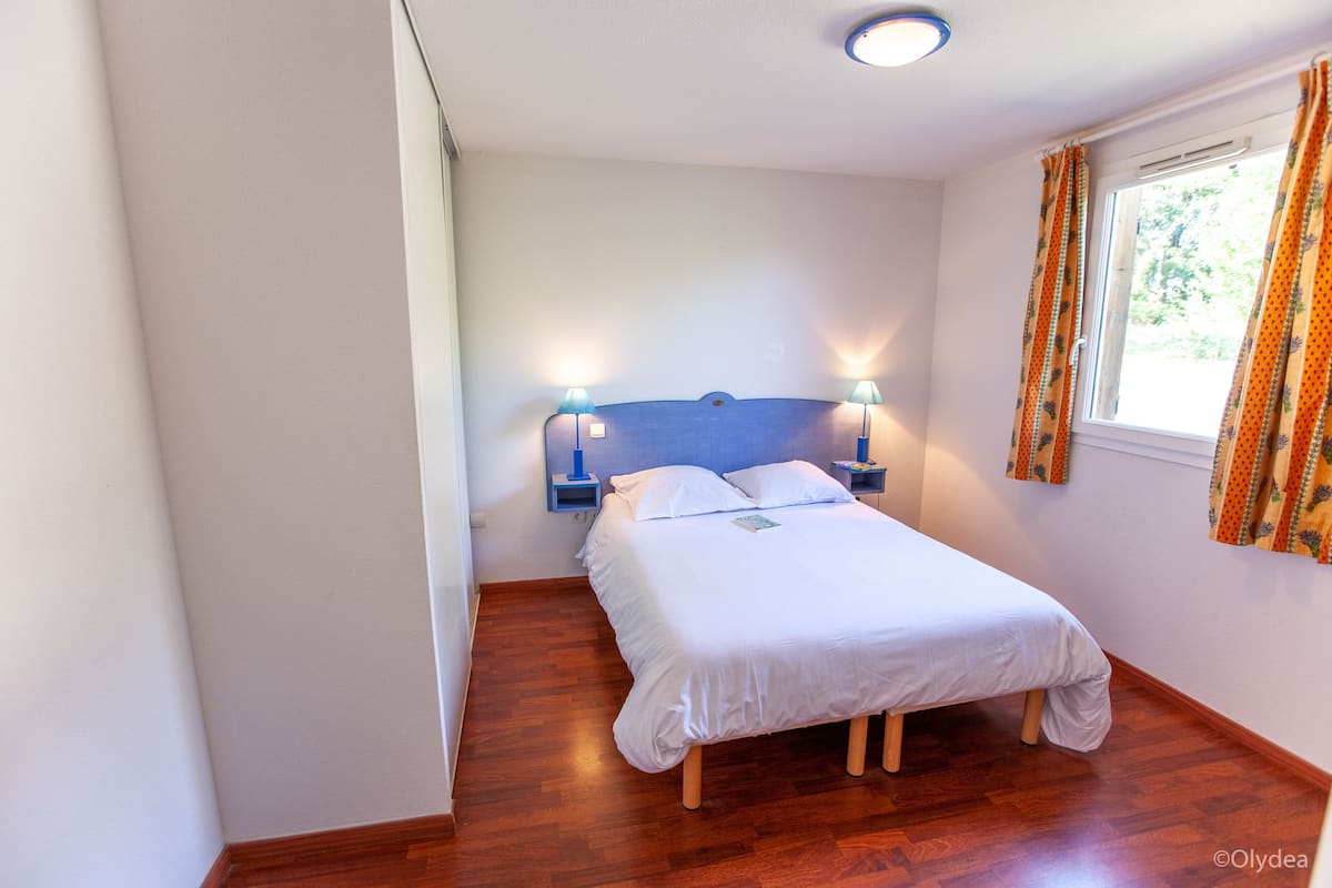 A small, neatly arranged bedroom with a single bed, two bedside lamps, and a window with curtains.