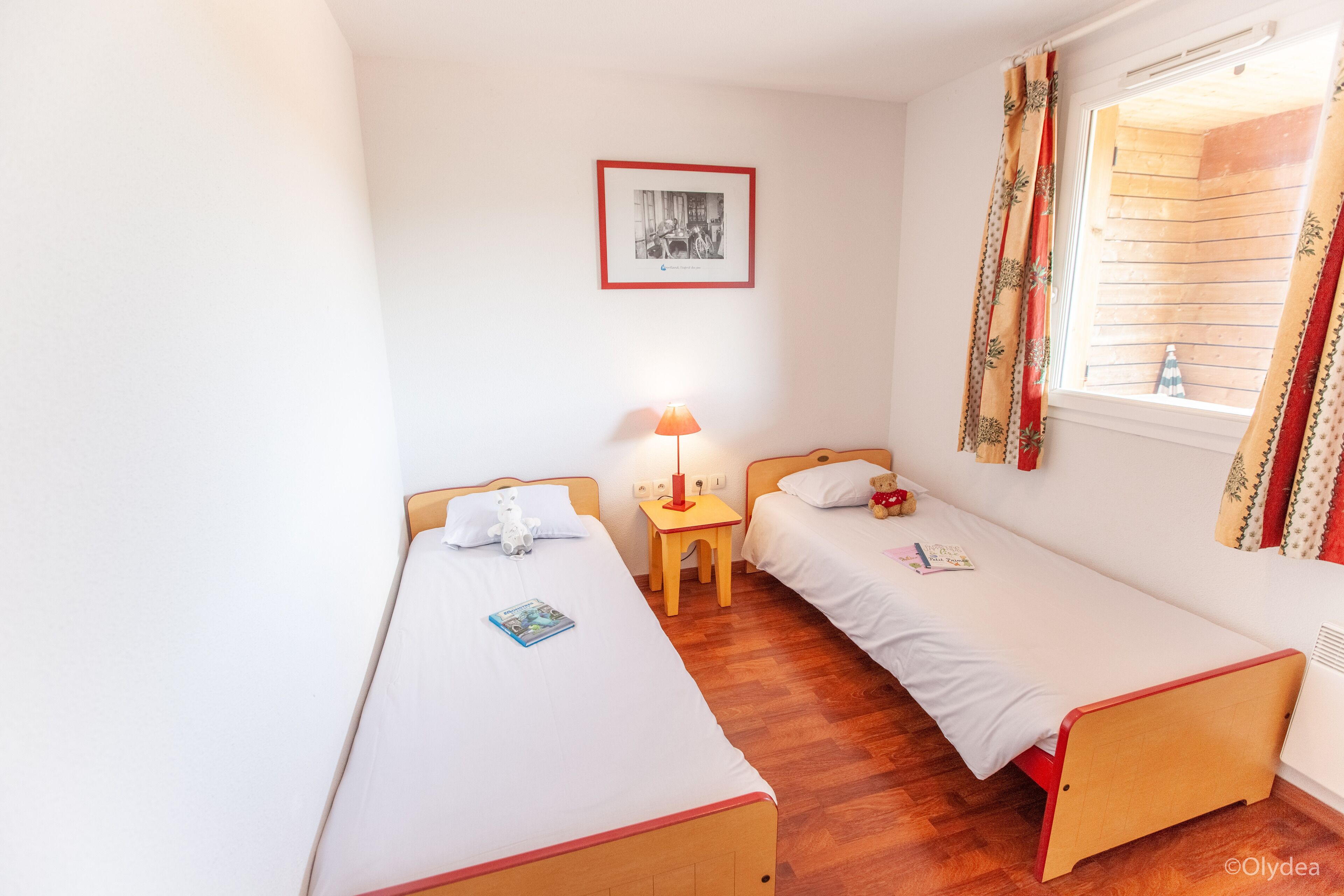 A small, clean room with two single beds, a nightstand with a lamp, a framed picture on the wall, and a window with curtains.