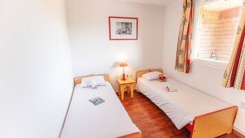 A small, clean room with two single beds, a nightstand with a lamp, a framed picture on the wall, and a window with curtains.