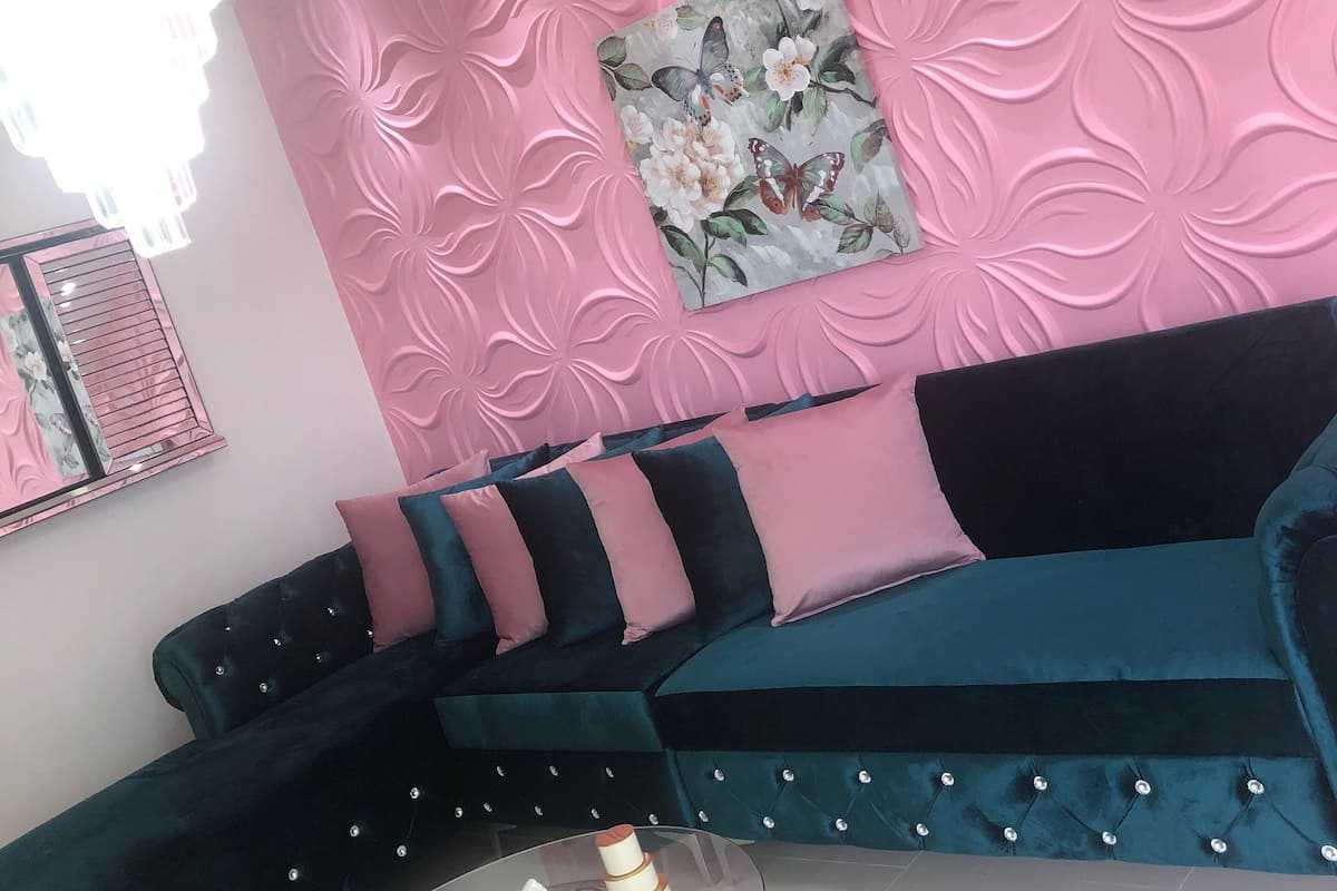 Un salon moderne avec un mur texturé de couleur rose, un tableau floral, un canapé bleu foncé agrémenté de coussins turquoise et une table basse en verre sur laquelle repose un objet blanc.