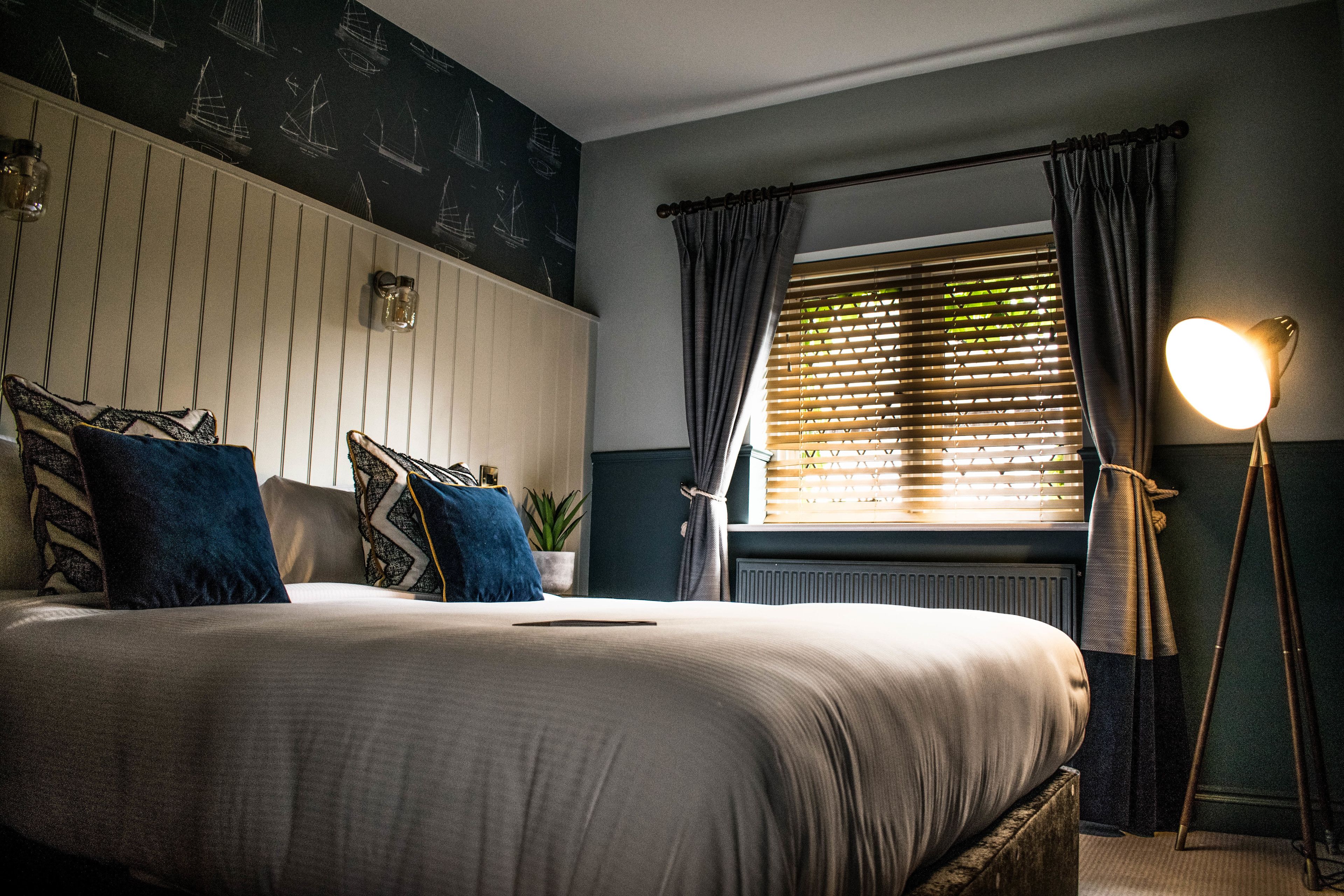 A neatly made bed with pillows, a bedside lamp, a window with curtains, and a wall with a nautical-themed mural.