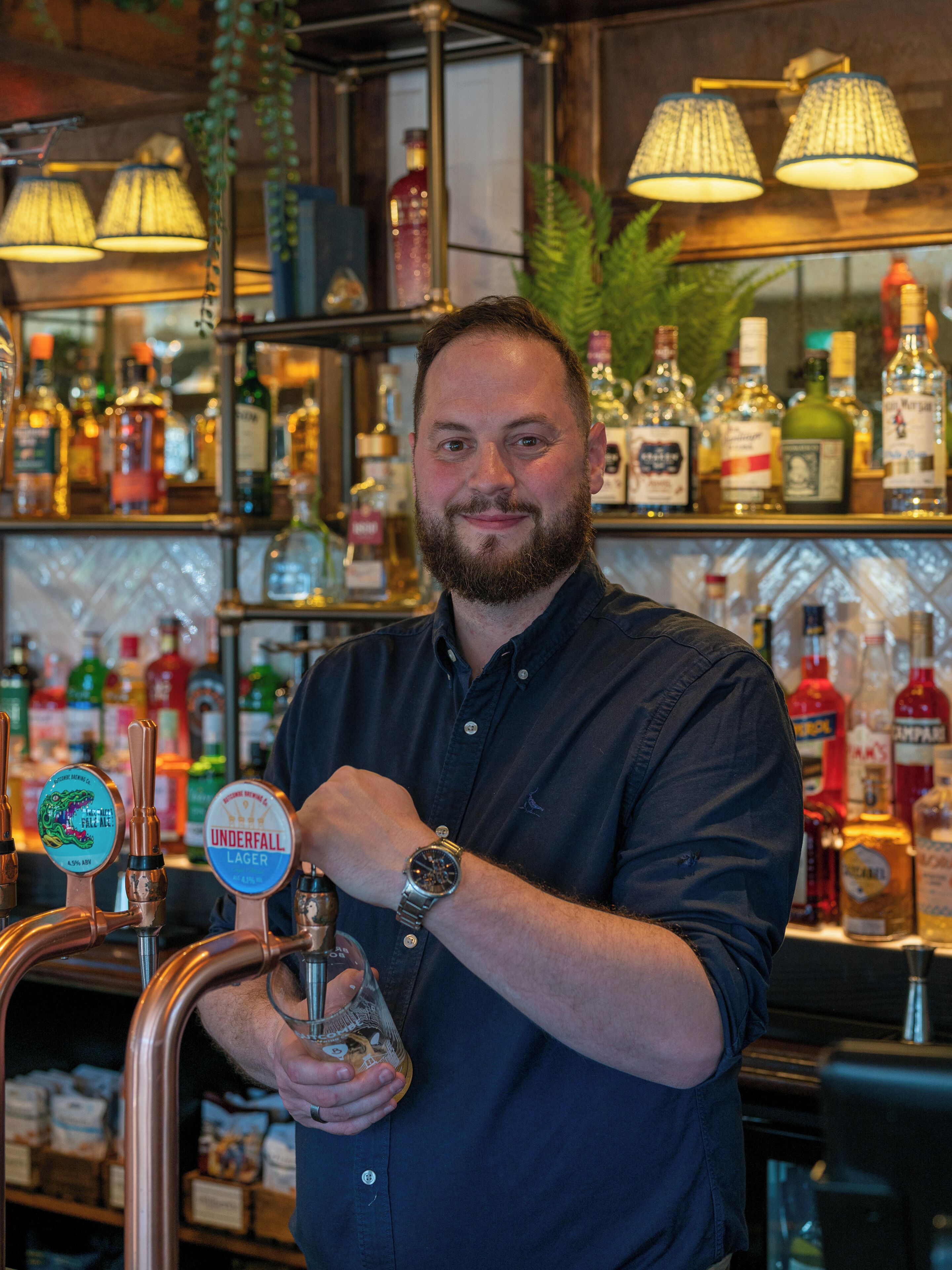 Una persona en un bar, con un grifo de cerveza que dice 'Underfall Lager' y varias botellas al fondo.