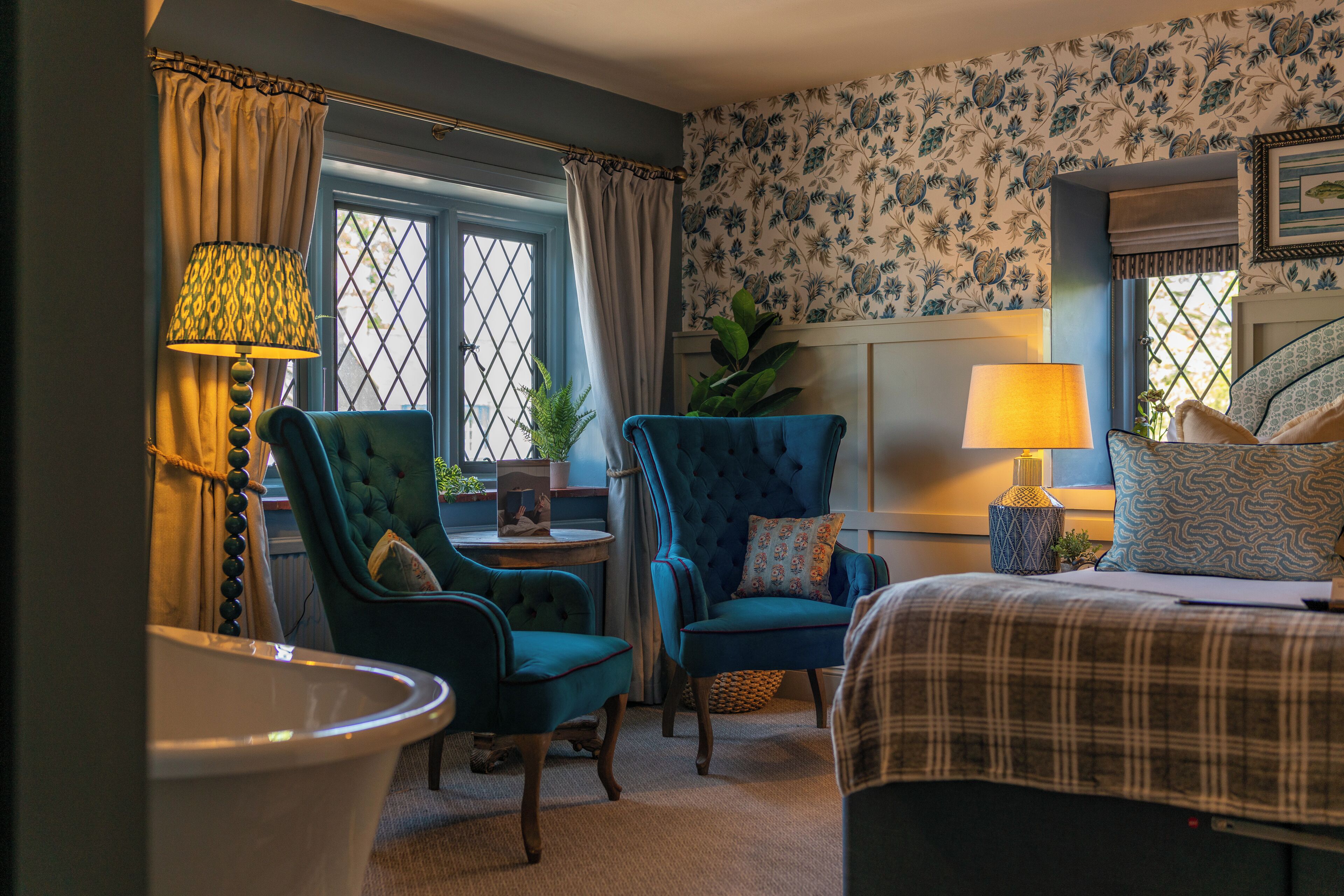 A bedroom with a bed, two chairs, a lamp, a window with curtains, and a wall with floral wallpaper.