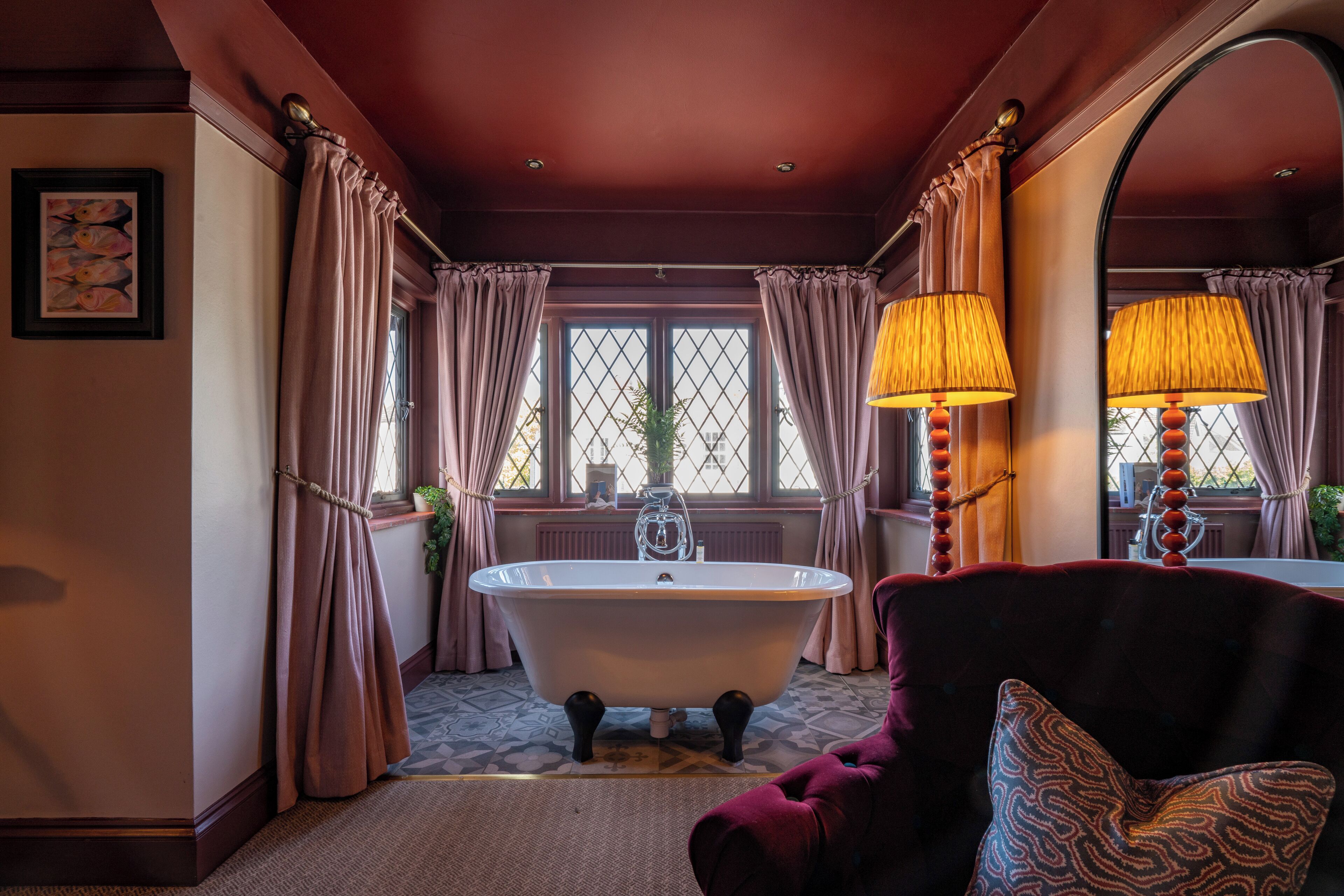 A bathroom with a freestanding bathtub, two lamps, a mirror, and a window with curtains.