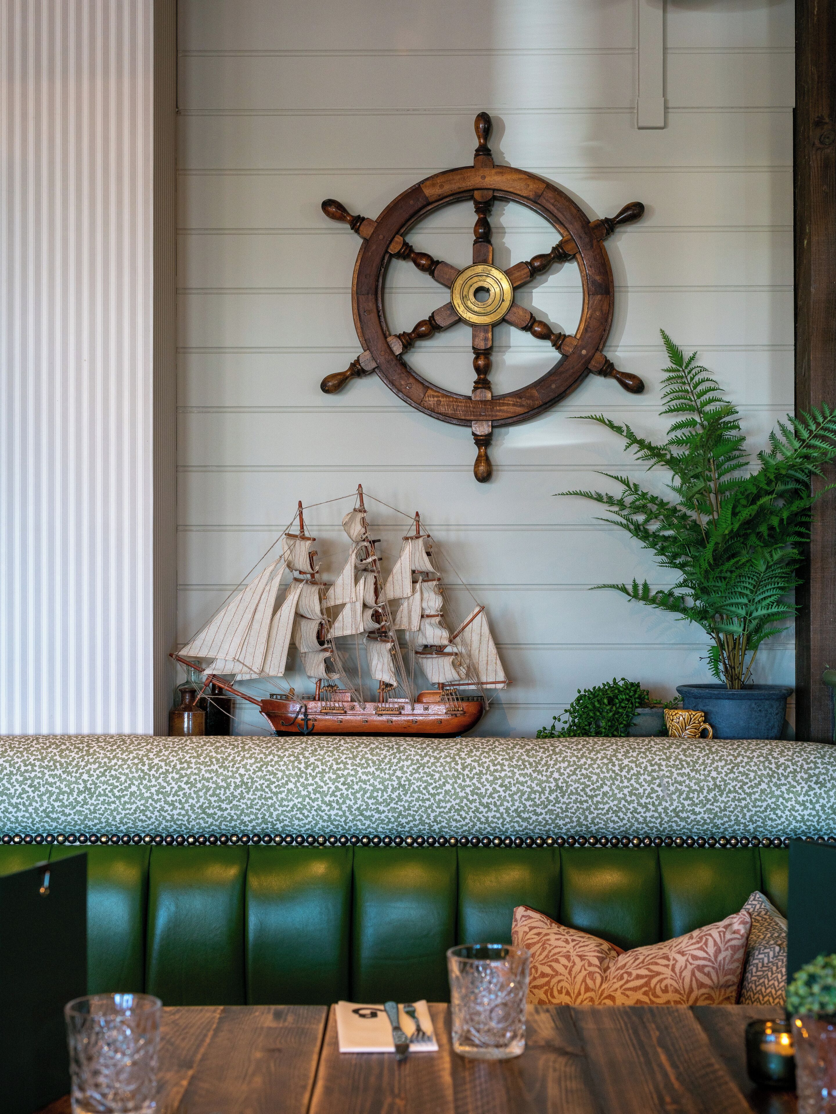 A ship's wheel, a model ship, a potted plant, a cushioned bench, and a table with a glass and cutlery.
