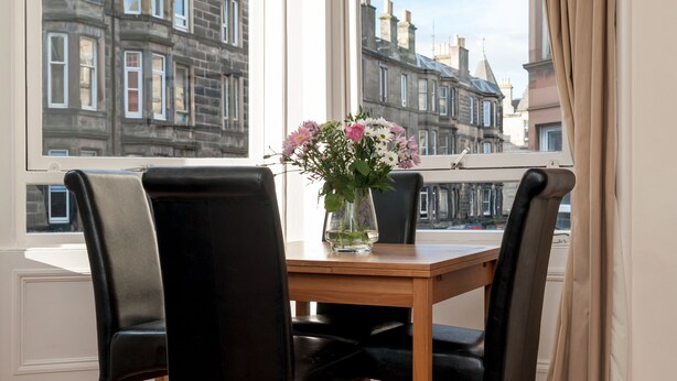 A dining table with four black chairs, a vase with flowers, and a view of historic buildings through large windows. - Edinburgh Rossie Apartment (Edinburgh)