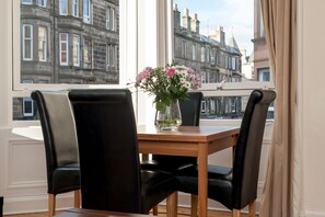 Una mesa de comedor con cuatro sillas negras, un jarrón con flores y vistas a edificios históricos a través de amplios ventanales.