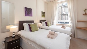 A hotel room with two beds, a nightstand, a lamp, and a window with curtains. - Edinburgh Rossie Apartment (Edinburgh)