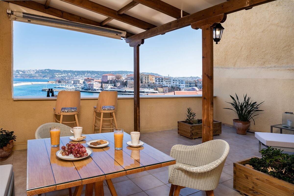 A terrace with a table set for breakfast, wicker chairs, potted plants, and a view of the sea and buildings.