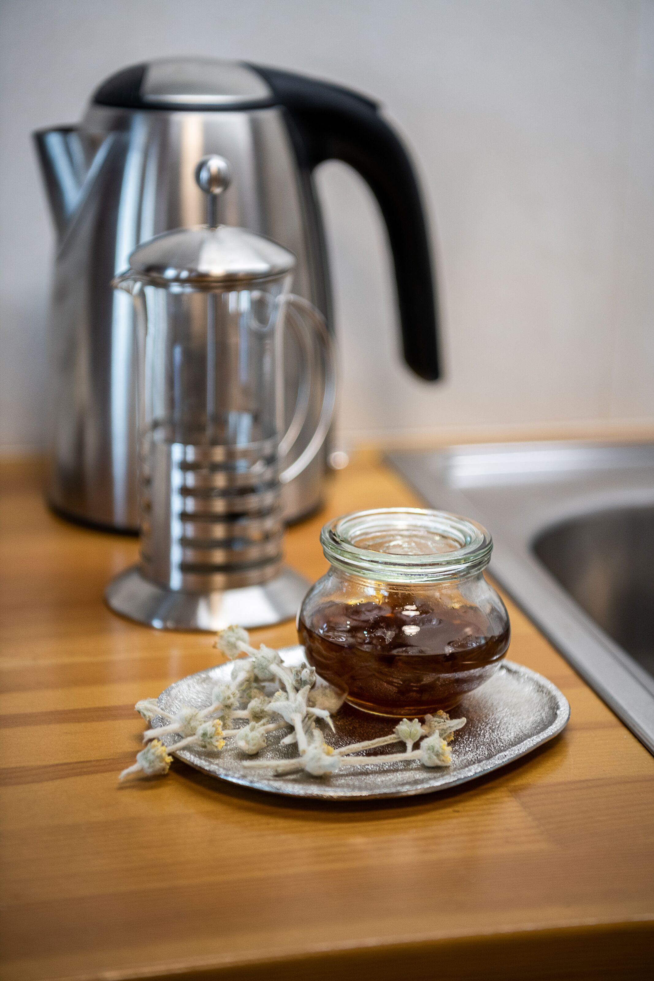A stainless steel kettle, a glass jar with a lid, and a plate with tea leaves on a wooden surface.
